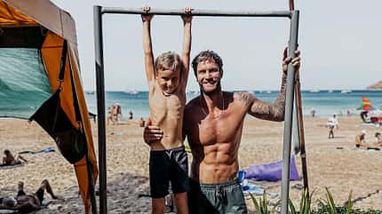 Matty Fox und sein Sohn an einem Strand. Der Kleine hängt an einer Klimmzugstange, während Matt ihn von der Seite hält. Matty Fox und sein Sohn an einem Strand. Der Kleine hängt an einer Klimmzugstange, während Matt ihn von der Seite hält.