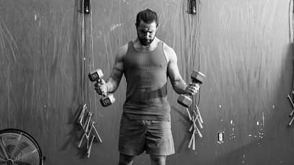 Ein Mann steht in einem Gym und hebt zwei Kurzhanteln. Er blickt konzentriert zu Boden. Ein Mann steht in einem Gym und hebt zwei Kurzhanteln. Er blickt konzentriert zu Boden.