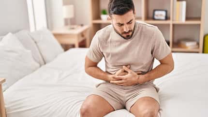 Ein Mann in beige gekleidet hält sich mit schmerzverzerrtem Gesicht den Bauch, während er auf einem weißen Bett sitzt. Ein Mann in beige gekleidet hält sich mit schmerzverzerrtem Gesicht den Bauch, während er auf einem weißen Bett sitzt.