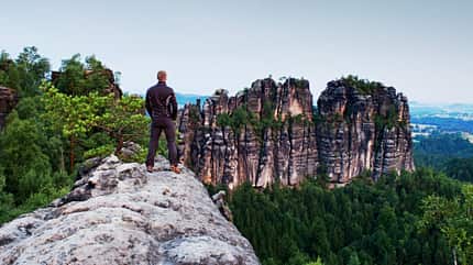 Rückansicht eines Mannes, der auf einem Berggipfel steht und auf eine Bergkulisse schaut Rückansicht eines Mannes, der auf einem Berggipfel steht und auf eine Bergkulisse schaut