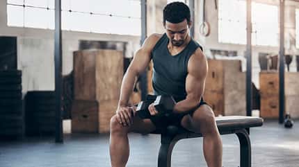 Aufnahme eines muskulösen jungen Mannes, der mit einer Hantel in einem Fitnessstudio trainiert Aufnahme eines muskulösen jungen Mannes, der mit einer Hantel in einem Fitnessstudio trainiert