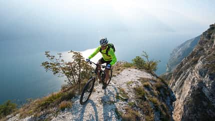 Extreme Sports,Men,Cycle,Sports Training,Insurance,Dirt Road,Fun,Mountain Bike,Action,Concentration,Happiness,Danger,Loneliness,Solitude,Risk,Speed,White,Outdoors,Athlete,Cycling,Activity,Italy,Sunlight,Rock - Object,Sun,Sky,Lake Garda,Lake,Fog,Water,Single Lane Road,Street,Free Ride,Trentino,Uphill mountainbiking over the lake garda