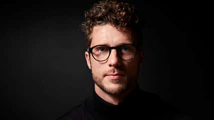 Ein Mann mit Locken und Brille vor dunklem Hintergrund