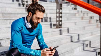 Mann mit Dutt sitzt auf der Treppe und schaut auf sein Smartphone Mann mit Dutt sitzt auf der Treppe und schaut auf sein Smartphone
