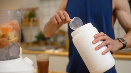 Man adding a scoop of chocolate protein powder into a blender filled with chopped fruits, preparing nutritious shake in the kitchen.