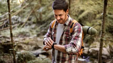 Mann mit Rucksack schaut beim Wandern im Wald auf seine Smartwatch.