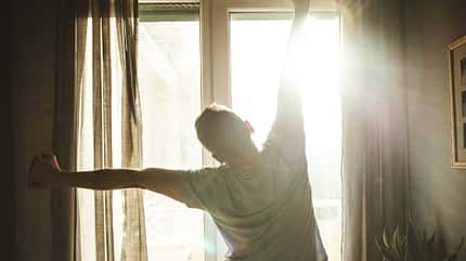Mann steht vor dem Fenster, durch das die Sonne scheint, und streckt sich