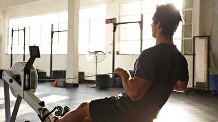 Mann trainiert im Fitnessstudio auf einem Ruderergometer und zieht den Griff zum Oberkörper.