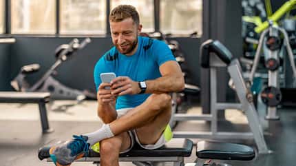 Sportlicher Mann mit Smartwatch am Handgelenk sitzt im Fitnessstudio auf einer Bank und schaut auf sein Smartphone.