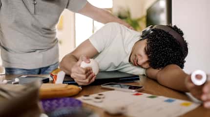Jugendlicher sitzt erschöpft über dem Tisch, während eine Person ihm beruhigend die Hand auf die Schulter legt.