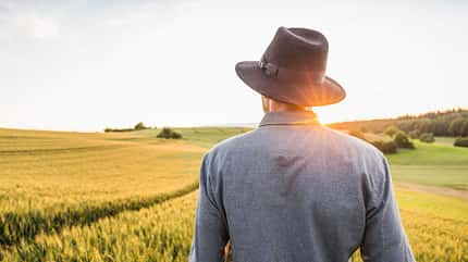 Ein junger Mann mit  Hut schaut über ein Feld, während die Sonne im Hintergrund untergeht.