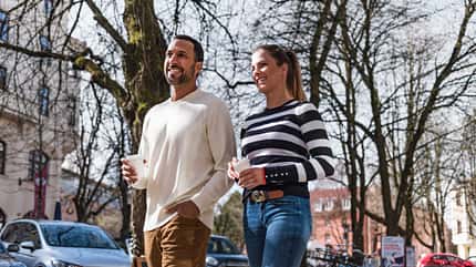 Ein junger Mann und eine junge Frau spazieren in einer städtischen Umgebung mit einem Kaffeebecher in der Hand.
