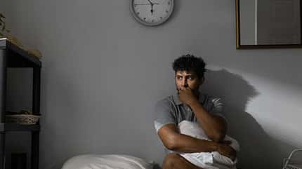 Ein Mann sitzt auf seinem Bett mit einem Kissen im Schoß und schaut nach draußen