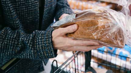Hände halten ein in Plastik eingepacktes Brot über dem Einkaufswagen im Supermarkt