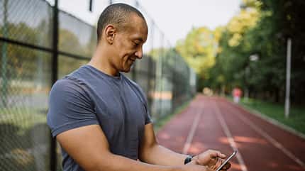 Ein Mann im T-Shirt steht draußen auf einer Laufbahn und schaut auf sein Smartphone Ein Mann im T-Shirt steht draußen auf einer Laufbahn und schaut auf sein Smartphone