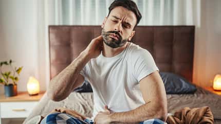 Mann in Schlafkleidung sitzt im Schneidersitz im Bett und fasst sich an den Nacken