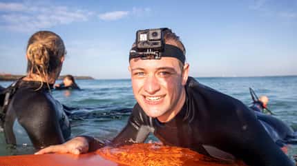 Junger Mann in Neoprenanzug und mit Actionkamera auf dem Kopf liegt im Wasser auf Surfbrett, Im Hintergrund andere Surfer im Meer