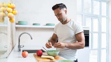 Ein junger Sportler schneidet sich in der Küche Obst in seinen Joghurt. Ein junger Sportler schneidet sich in der Küche Obst in seinen Joghurt.