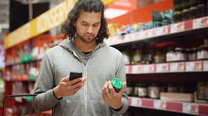 Mann steht im Supermarkt und hält in der einen Hand sein Smartphone, in der anderen ein Konservenglas Mann steht im Supermarkt und hält in der einen Hand sein Smartphone, in der anderen ein Konservenglas