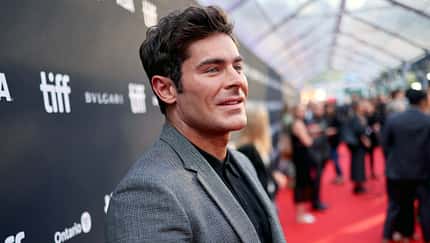 Zac Efron steht in grauem Anzug bei einer Filpremiere vor einer Werbewand.