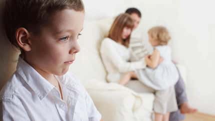 Mutter, Vater und Tochter beschäftigen sich auf dem Sofa, während der Sohn in der Ecke steht und schmollt