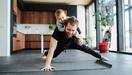 Ein Vater macht Liegestütze in der Küche während sich sein Baby auf seinem Rücken festhält