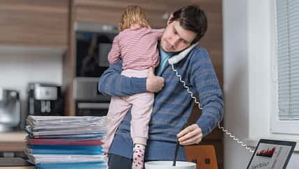 Ein Vater beim Multitasking in der Küche mit seinem Kind auf dem Arm und einem Telefonhörer unters Kinn geklemmt