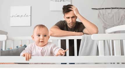 Baby lugt übers Kinderbettchen, der Vater steht dahinter mit einem verzweifelten Blick