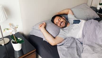 Weißer, dunkelhaariger Mann liegt in einem grauen Bett, streckt sich und lächelt dabei