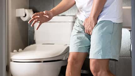 Ein weißer Mann in heller Kleidung mit kurzer Hose steht in einem Badezimmer und hält sich den Schritt durch dringenden Harndrang