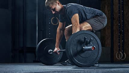 Ein weißer Mann mit dunklen Haaren und in Sportkleidung gekleidet, befindet sich in einem Fitnessstudio und führt eine Übung mit einer Langhantel durch