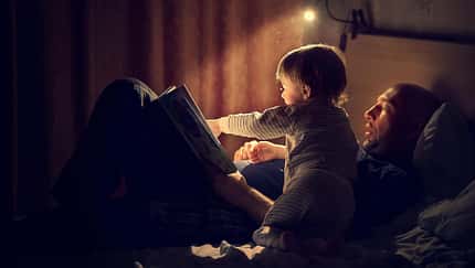 Ein Vater liegt mit seinem Kind im Bett und liest ihm aus einem Buch vor
