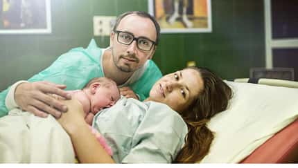 Eine Wöchnerin liegt mit ihrem Baby auf dem Bauch in einem Krankenhausbett, der Vater des Kindes sitzt daneben und umarmt die beiden