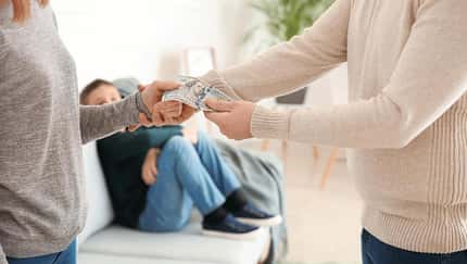 Ein Mann drückt einer Frau ein paar Geldscheine in die Hand. Im Hintergrund sieht man ein Kind, das auf dem Sofa sitzt