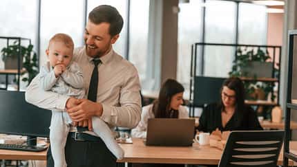 Ein Vater im Business-Outfit hält in einem Büro ein kleines Kind auf dem Arm, im Hintergrund sieht man Kolleginnen bei der Arbeit