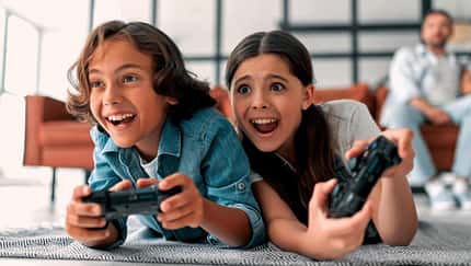 Man sieht zwei Kinder auf dem Teppich liegend auf einer Konsole spielen. Im Hintergrund sitzt der Vater auf dem Sofa.