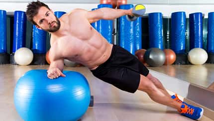 Mann macht Seitplanke auf Gymnastikball und dabei Seitheben mit einer Kurzhantel.