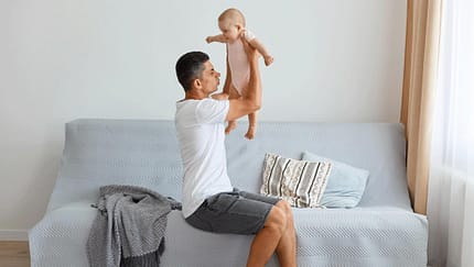 Ein Vater sitzt auf dem Sofa, hält sein Baby in die Höhe und riecht an der Windel