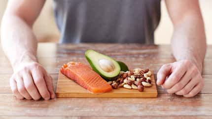 Junger Mann sitzt am Tisch, auf dem ein Brett mit Lachs, Avocado und Nüssen liegt.