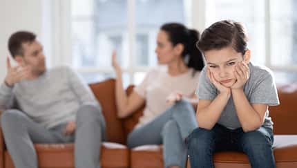 Man sieht im Vordergrund einen kleinen Jungen, der genervt guckt, im Hintergrund sitzen seine Mutter und sein Vater auf dem Sofa und diskutieren.