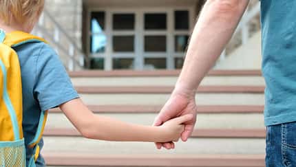 Man sieht in einer Nahaufnahme Vater und Sohn von hinten Hand in Hand zur Schule gehen.