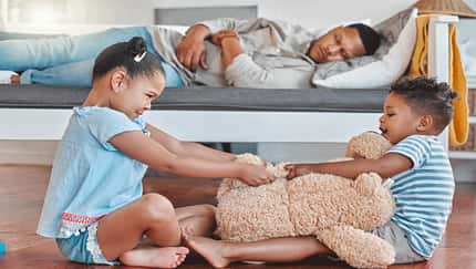 Zwei Kinder streiten sich im Wohnzimmer, dahinter liegt der Vater erschöpft auf einer Couch
