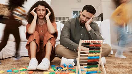 Eine Mutter und ein Vater sitzen verzweifelt auf dem Boden des Kinderzimmers, ihre beiden Kinder toben um die beiden herum