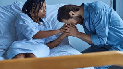 Eine Frau liegt in einem Krankenhausbett, ein Mann sitzt gekrümmt davor und weint.