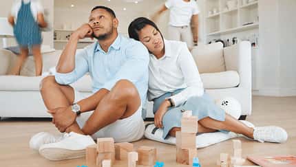 Vater und Mutter sitzen erschöpft vor dem Sofa, im Hintergrund sieht man zwei Kinder fröhlich auf der Couch hüpfen