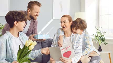 Eine Frau sitzt auf dem Sofa, ihr Mann und ihre Kinder reichen ihr Blumen und Schokolade und eine Karte zum Muttertag
