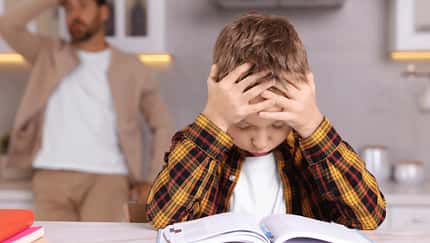 Ein kleiner Junge sitzt angestrengt über einem Schulbuch, im Hintergrund sieht man einen verzweifelten Vater