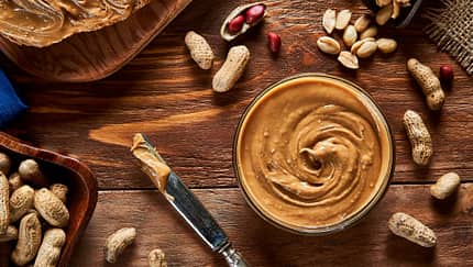 Erdnussbutter in einem Glas auf Holzuntergrund mit einem Messer Erdnussbutter daneben und Erdnüssen