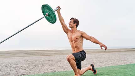 Side view of a young muscular man training on the beach shirtless doing landmine overhead press with barbell in kneeling position.