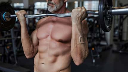 Ein Mann mit Sixpack trainiert oberkörperfrei seinen Bizeps mit einer Langhantel